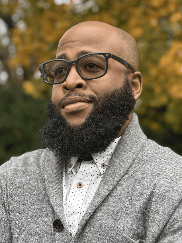 L'Rae Brantley in grey shawl neck sweater and white and black polka dot button down shirt.